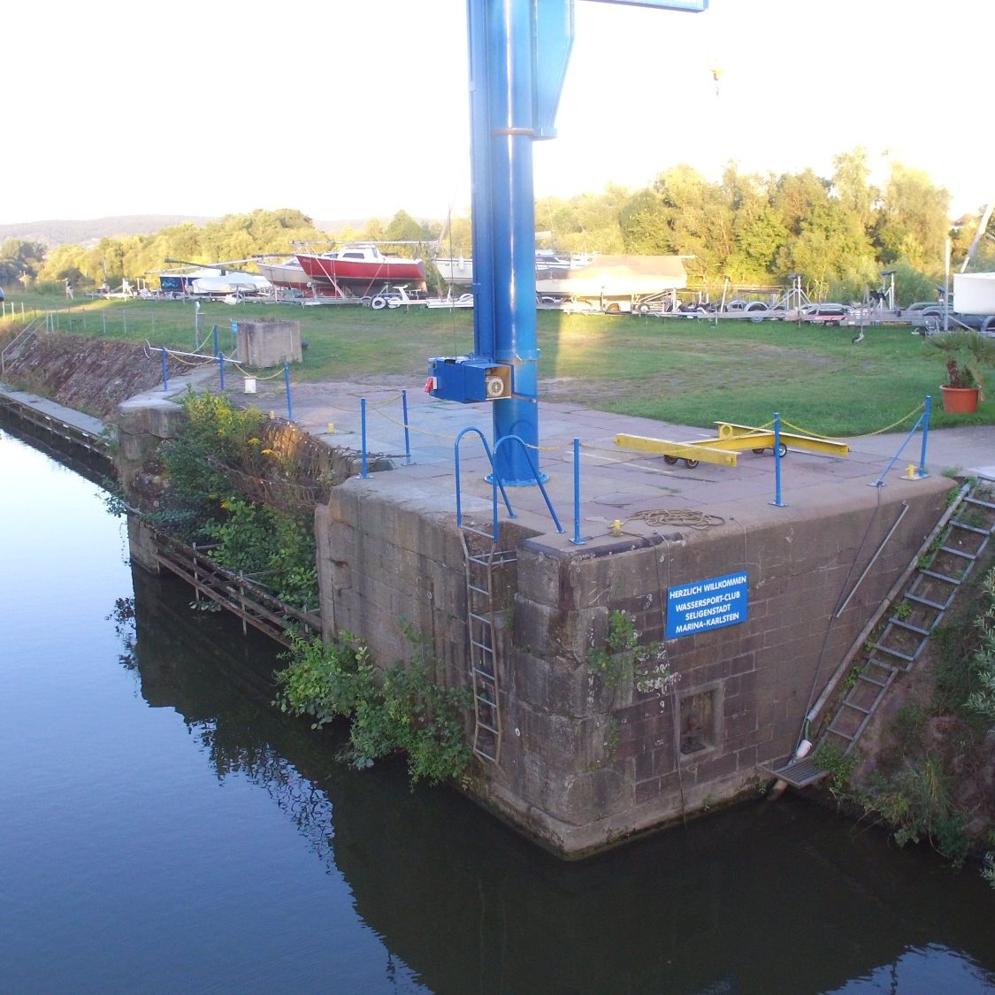 Hafenanlage Hafenanlage mit Kran und Steg am Wasser, umgeben von Bäumen und Booten.